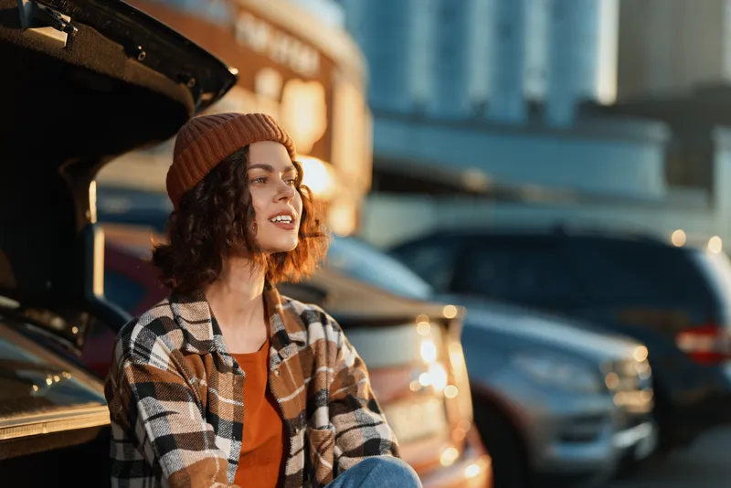 woman car trunk parking smile beanie candid urban Young woman sits at an open car trunk in a city parking lot during golden hour glow, authenticity and mindful living captured with emotional