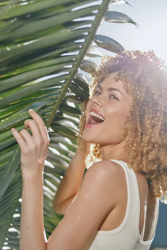 smiling woman with curly hair enjoys tropical sunlight, holding palm leaves, wearing a white tank top in outdoor summer setting on bright day