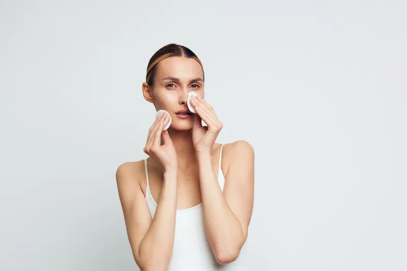 A young woman with clear skin removes makeup using cotton pads on her face. She wears a white tank top against a plain white background. Concept for skincare and beauty routine.