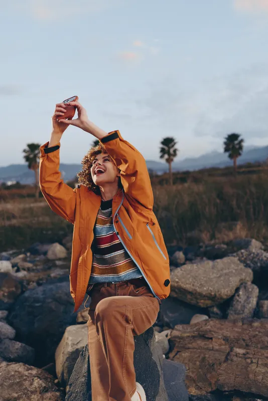 Cheerful woman outdoors, orange jacket, hands near face in joyful smile pose, hiking on rocks trail, soft breeze, distant mountains, palms and cloudy sky, lifestyle moment