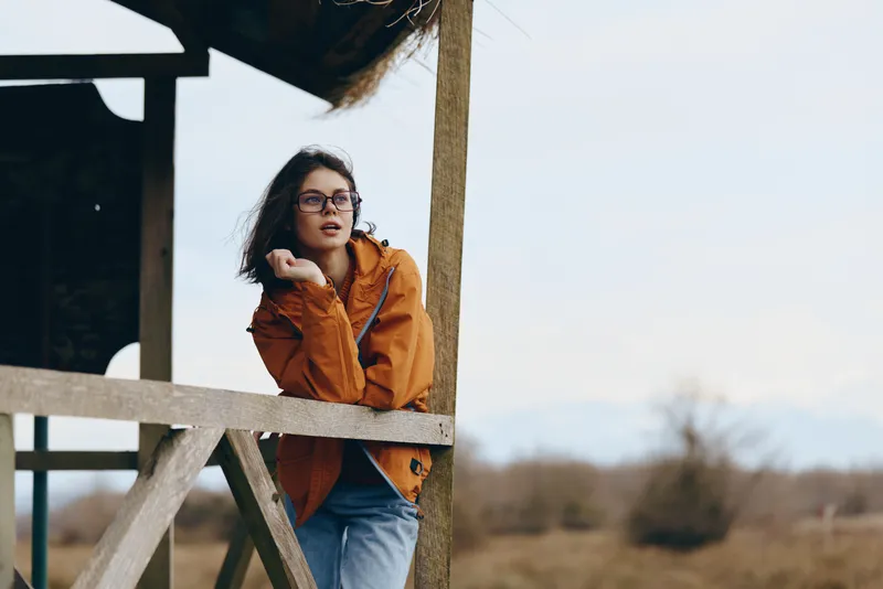 Woman outdoors lifestyle orange jacket glasses wind portrait spring nature, relaxed pose through wooden frame, soft daylight, hair blowing, casual photo, rural landscape