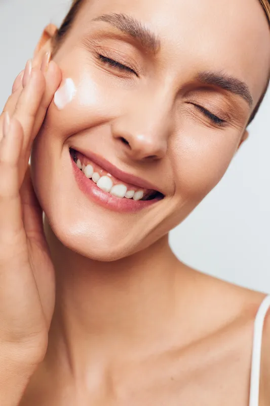 Smiling woman applies moisturizing cream on face with closed eyes shows healthy skin and skincare routine isolated on white background closeup portrait beauty concept.