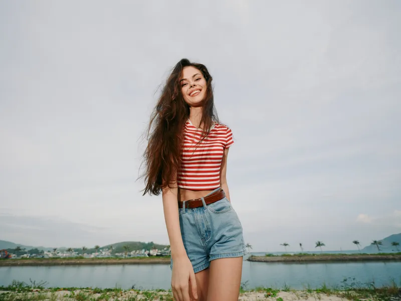 A young woman poses outdoors in a casual summer outfit, striped crop top and denim shorts, with a confident smile and relaxed stance under a bright open sky.