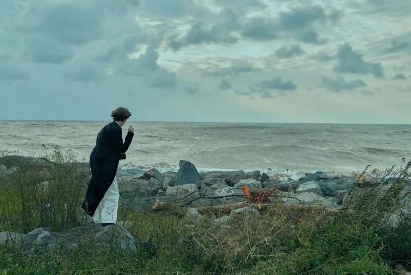 Isolation, contemplation, seascape, shoreline scene with a figure in a dark coat gazing at the ocean under a moody sky, evoking reflection and calm moments