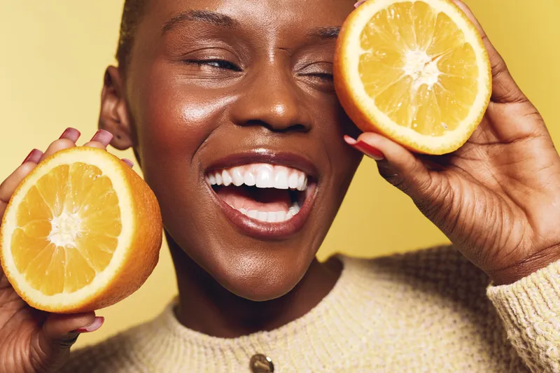 Happy black woman with dark skin holds two orange halves near face smiling brightly on yellow background. Concept of healthy lifestyle, vitamin c, natural beauty and wellness.