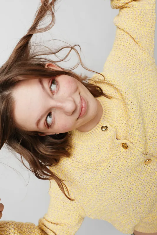 Young woman with long brown hair wearing a yellow knitted sweater looks sideways with a playful smile. Studio portrait on a light background with natural expression.