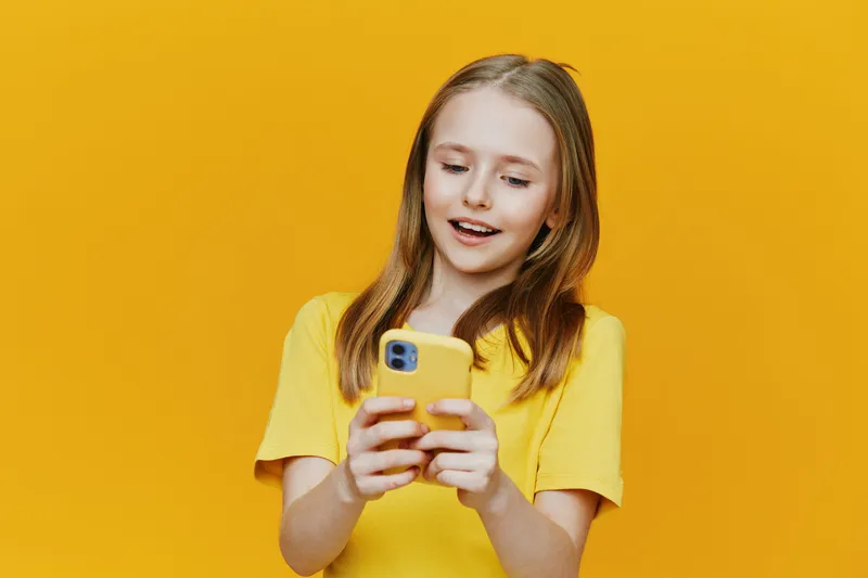Happy girl using smartphone with yellow case, dressed in bright yellow t shirt, against vibrant yellow background, expressing joy and excitement, youthful emotion