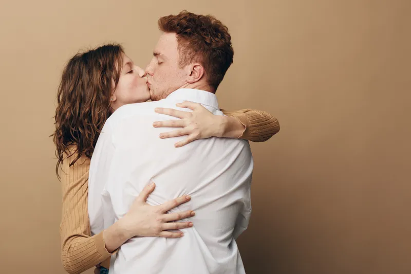 Young couple embraces and kisses tenderly against beige background. Woman with curly hair hugs man in white shirt. Concept of love, affection, romance, and intimate relationship.