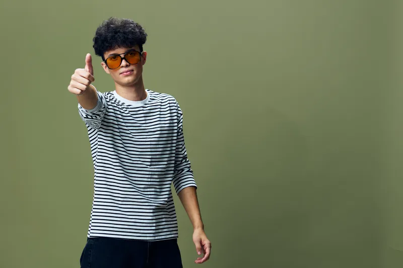 Young man wearing striped shirt and orange sunglasses gives thumbs up against green background, smiling and confident with casual style and relaxed posture.