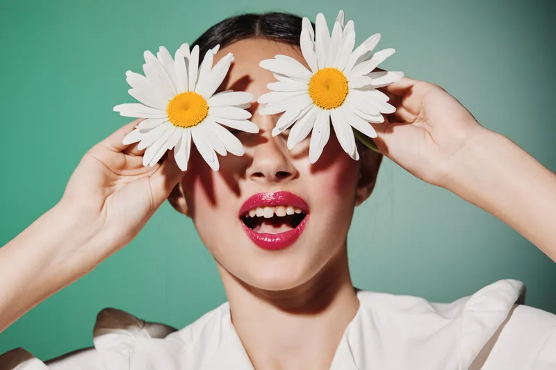 Young woman holding two daisies in front of her eyes with bright makeup and red lipstick on a green background. Fashion creative cosmetic concept focused on beauty and playfulness.