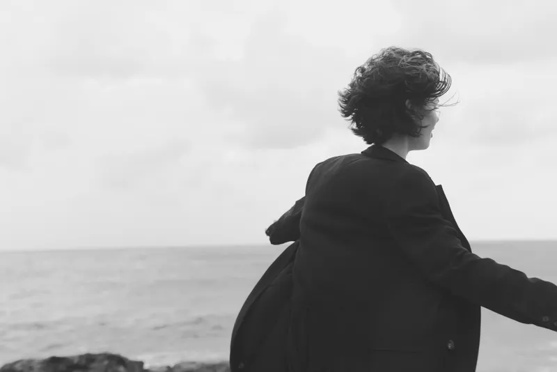 Figure in a dark coat stands at the wind swept coast, back to the camera, facing the sea and horizon, suggesting contemplation and quiet courage in an overcast scene