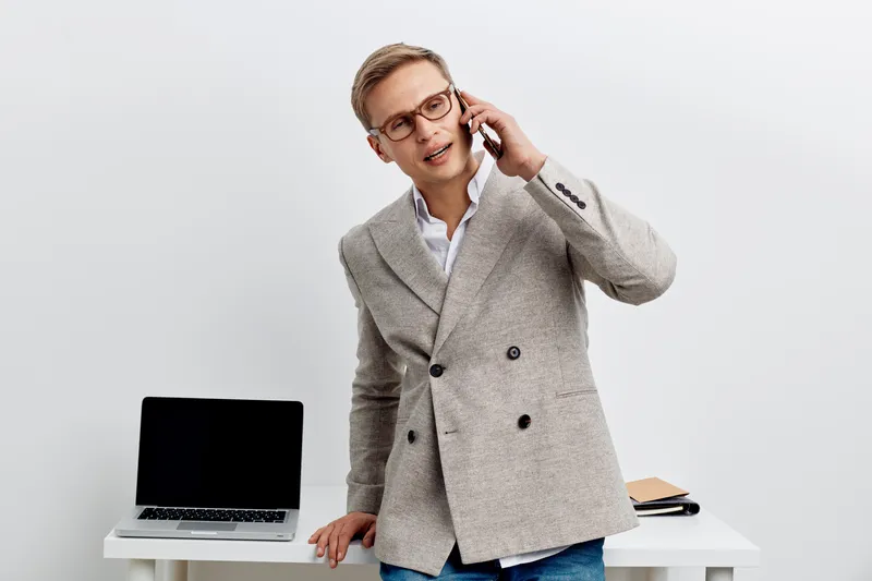 young caucasian man working smart casual jacket talking smartphone standing near laptop white background digital nomad concept