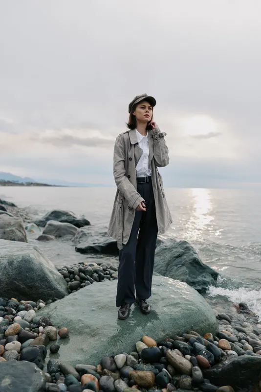 Stylish woman in long coat and hat stands on rocky shore near calm sea under cloudy sky. Moody natural light highlights fashion and contemplative mood outdoors.