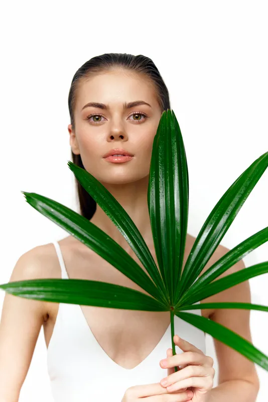 Спокойная молодая женщина неопределенной этнической принадлежности держит ярко-зеленый пальмовый лист, воплощая естественную красоту на фоне чистого белого фона