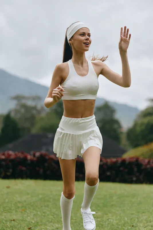 Young athletic woman running outdoors in a white sports outfit, showcasing fitness and vitality in a natural setting with greenery and mountains