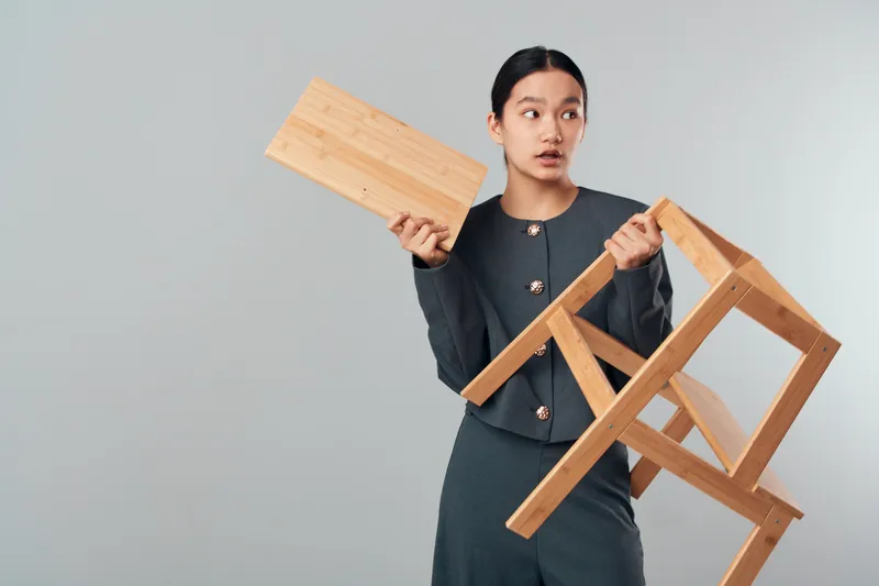 Woman holding wooden chair and board looking aside with surprised expression on gray background. Female in dark outfit posing with furniture pieces for design and creativity idea.