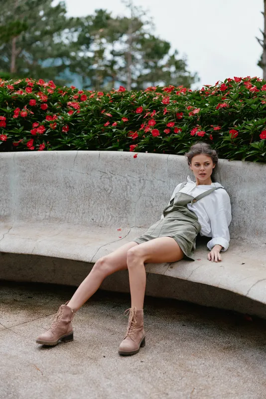 Casual summer fashion portrait of a young woman in overalls, sitting elegantly in a garden surrounded by vibrant flowers, showcasing a relaxed and graceful mood