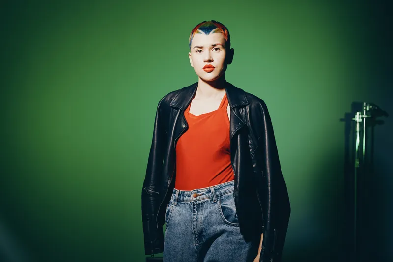 young woman with colorful hair wearing a black leather jacket and orange top standing against a green background, casual style and modern fashion outfit