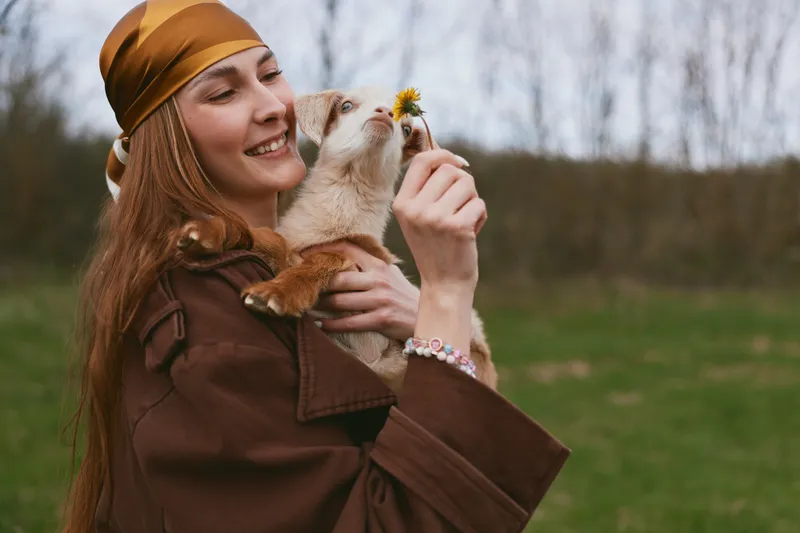 Woman holding a goat kid in the countryside, outdoor moment with smile and affection, nature colors, gentle breeze, rustic jacket, candid lifestyle