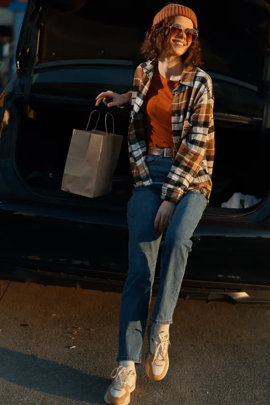 Woman sitting on car trunk with shopping paperbag, casual outfit and smile in sunlight golden hour glow; candid lifestyle portrait highlighting authenticity, mindful living and emotional storytelling.