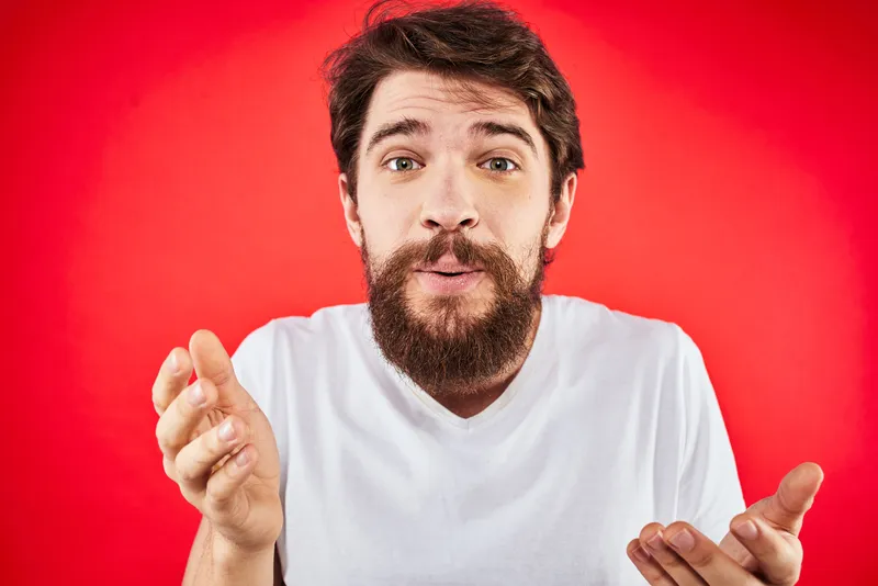 Бородатый мужчина, выражающий веселые эмоции, образ жизни, обрезанный вид, белая футболка, красный фон