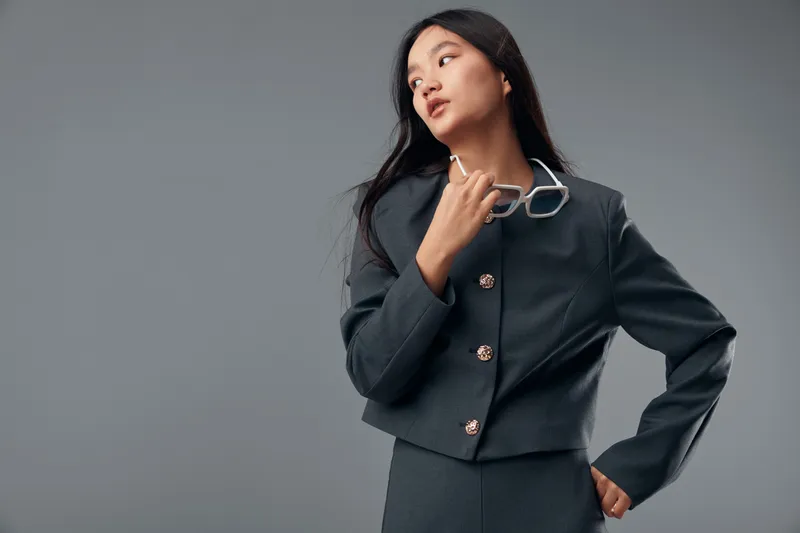Young woman in stylish dark grey blazer and matching skirt posing with white glasses in hand looking away on grey background. Fashion model with confident attitude and long hair in studio.