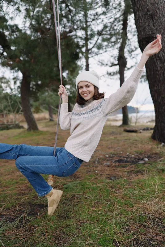 Woman on a swing in a park wearing a knitted sweater and beanie, enjoying a carefree moment in nature on a chilly day.