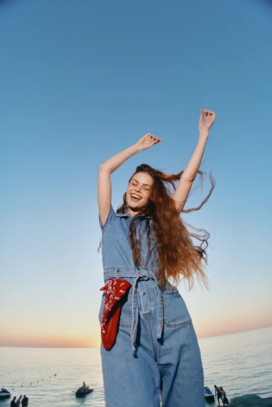 Lifestyle woman in modern boho-western denim street style joyfully dancing outdoors with film color effect and long flowing hair under clear sky at sunset.