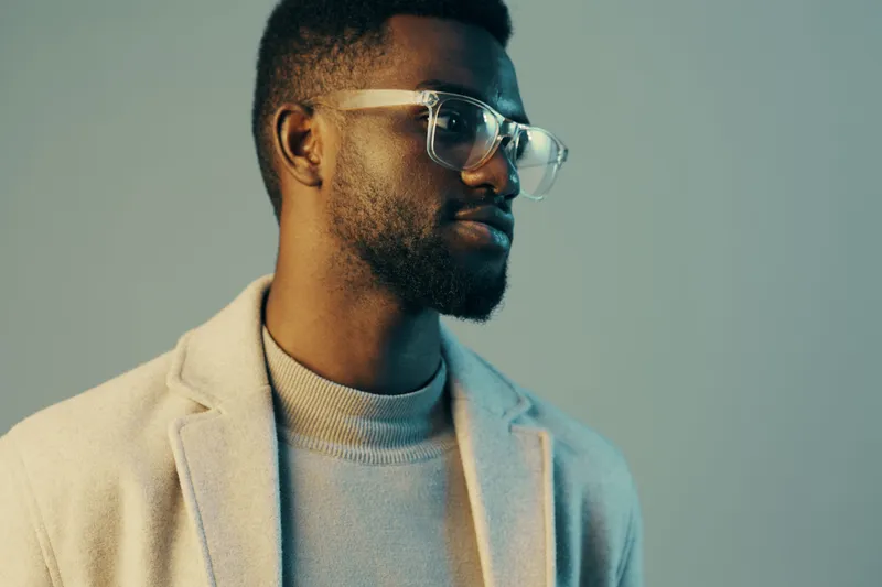 Stylish young Black man in clear glasses, wearing a light sweater and coat, showcasing a confident expression against a soft blue background