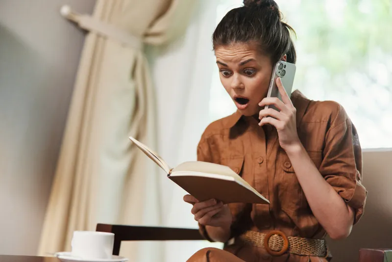 Потрясённая молодая женщина в коричневом наряде читает книгу и одновременно разговаривает по телефону, создавая эмоциональный момент удивления и любопытства на мягком нейтральном фоне