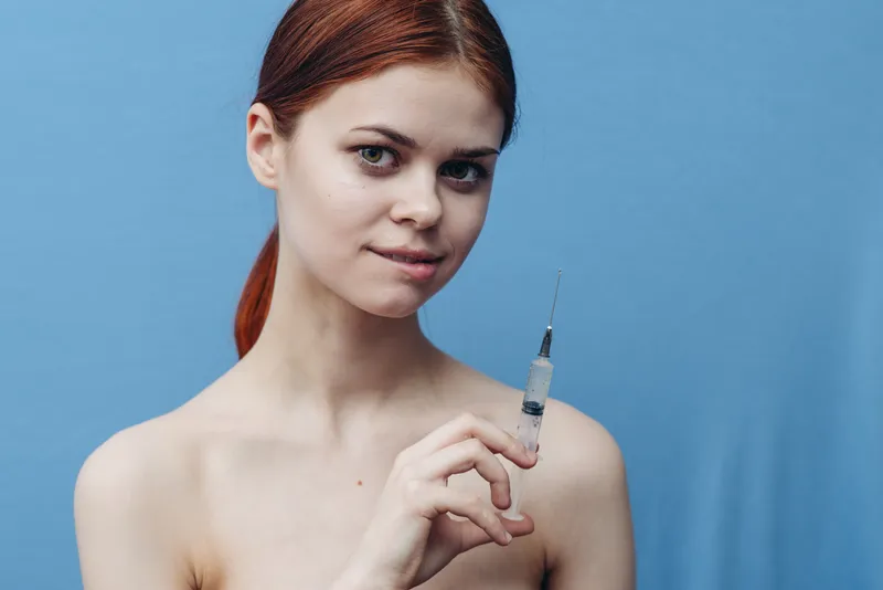 Женщина на синем фоне держит в руке шприц, обнаженные плечи, инъекция ботокса