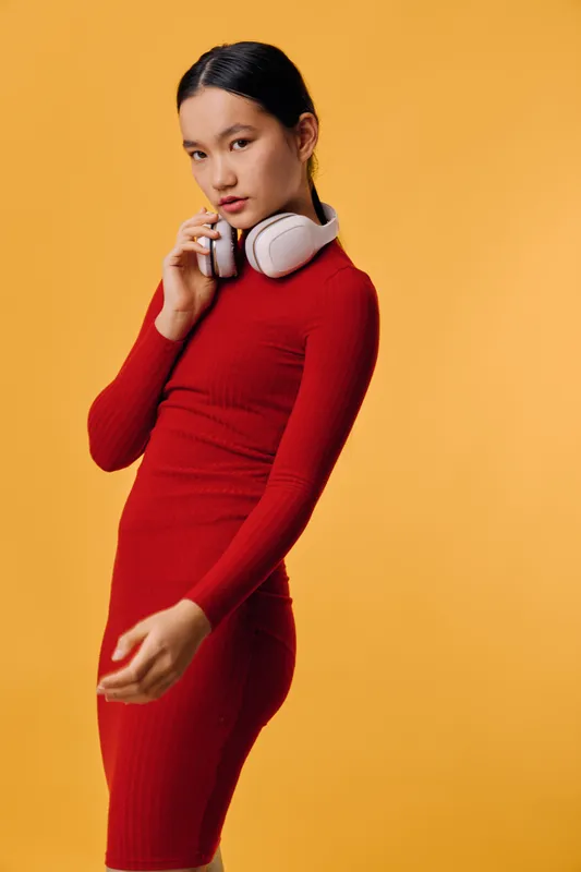 Woman in red dress with white headphones on orange background. Portrait of young female with black hair styled back, showing thoughtful expression and casual pose in studio lighting.