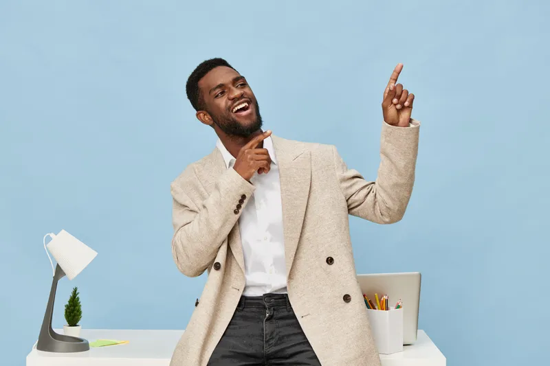 Enthusiastic smiling man in smart casual attire standing in modern office, gesturing with his finger, confident expression, blue background, workspace with computer and stationery, professional lifest