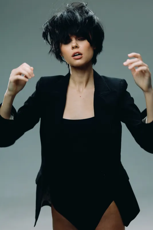 Stylish woman with black messy hair wearing a black blazer and dress posing against gray background.