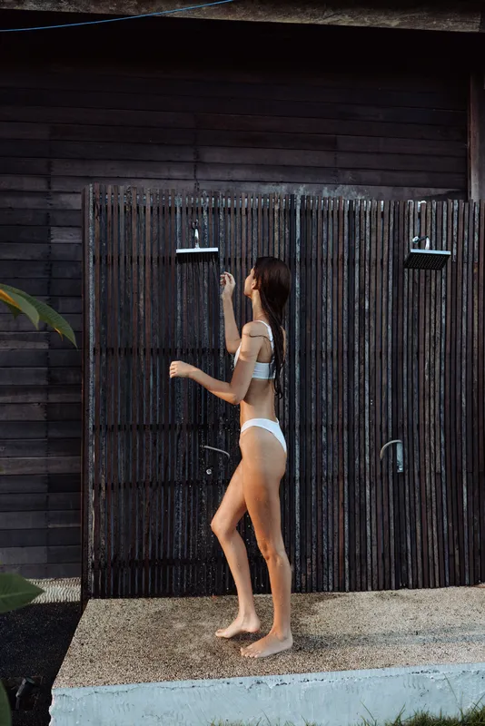 A woman in a white swimsuit walks beside an outdoor shower in a sunny setting, embracing a casual lifestyle moment. The scene captures everyday relaxation, personal care, and outdoor leisure,