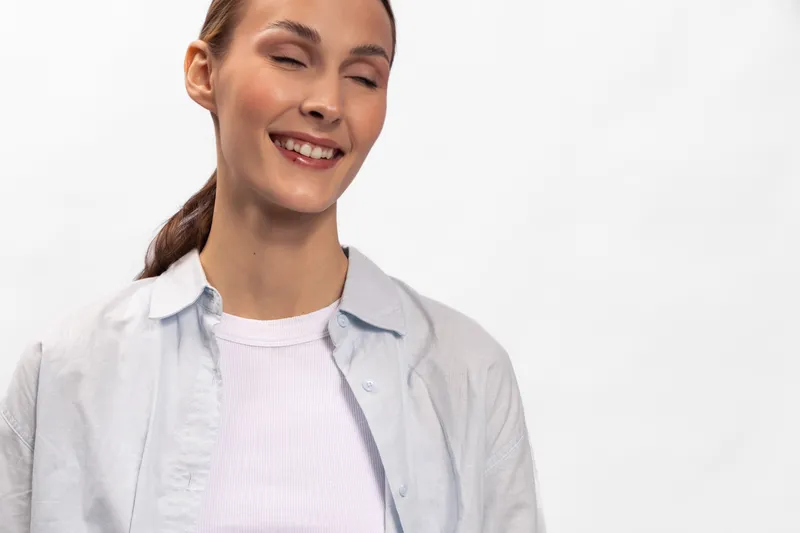 Smiling young woman with closed eyes wears casual white shirt and sweater, isolated on white background. Concept of happiness, relaxation, and natural beauty in studio.