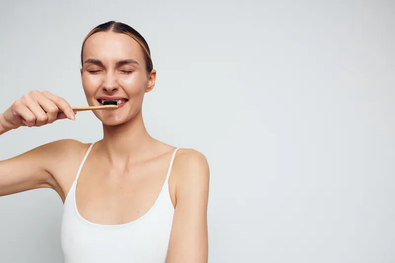 A young woman with closed eyes brushes her teeth using a bamboo toothbrush against a plain light background. Concept for dental hygiene, eco friendly lifestyle, and self care.
