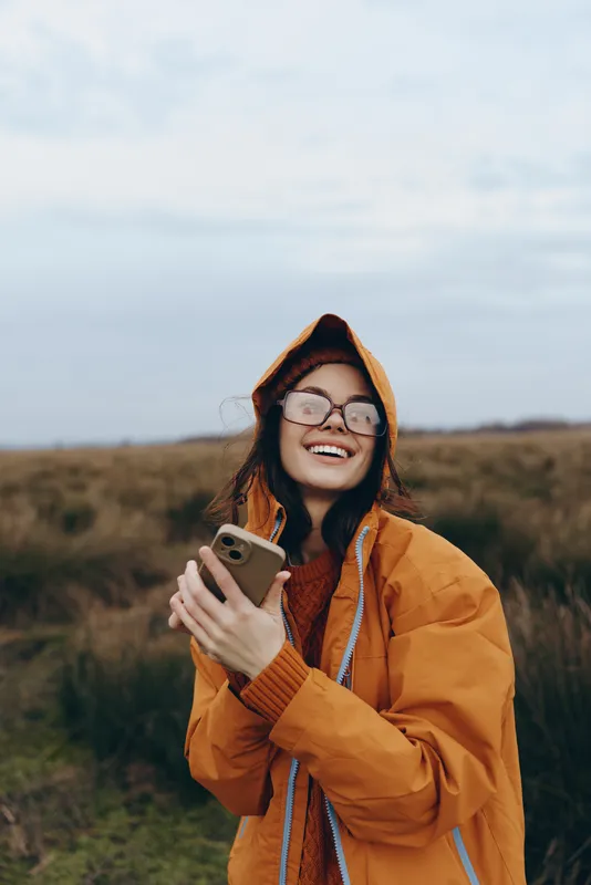 Woman lifestyle selfie smartphone outdoors field autumn hood glasses jacket, smiling and holding a phone, casual jacket and scarf, natural daylight in open countryside, travel moment