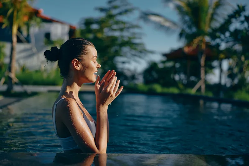A young woman in a white swimsuit relaxes in an outdoor pool with palms and a modern building in the background, enjoying a peaceful lifestyle moment on a sunny day.