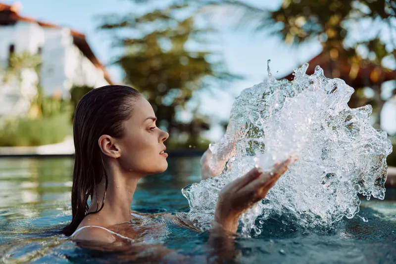 A relaxed woman enjoys a sunny pool day, water splashes around as she gazes upward, embodying a lifestyle moment of leisure, self care, and outdoor refreshment.