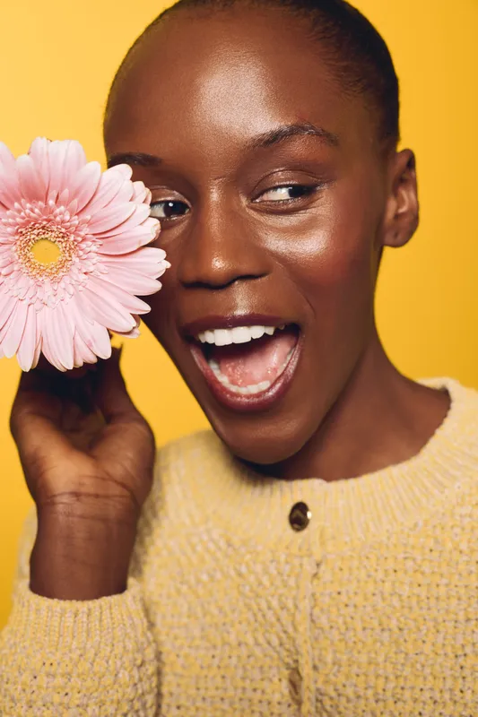 Smiling dark skinned African woman holds pink flower near face, wearing yellow sweater. Studio portrait on yellow background. Concept of beauty, happiness, skincare, and natural glow.