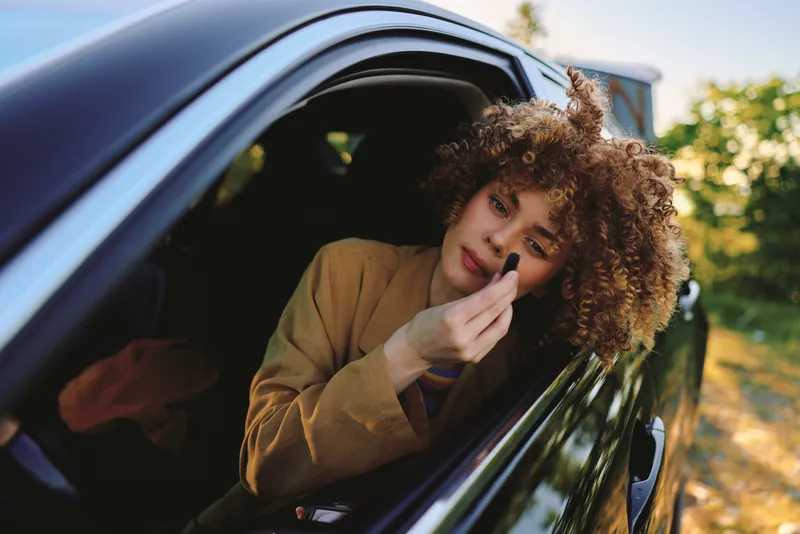 A young woman with curly hair sits in a car and applies lipstick through the open window, capturing a candid moment of casual beauty during a sunlit outdoor drive.