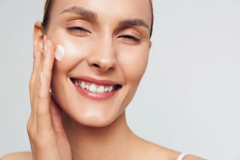 Smiling woman applying white facial cream on her cheek. Close-up on a light background. Concept of skincare, wellness, and self-care. Soft studio lighting creates a warm, fresh mood.