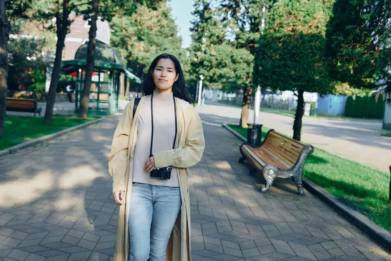 Woman walking in a park wearing a long beige coat and jeans, sunlight filters through trees creating a calm outdoor scene with benches and pavement
