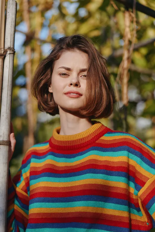 Woman portrait in colorful striped sweater outdoors, young woman with bob haircut and soft smile, holding playground pole, natural light and shallow depth of field.