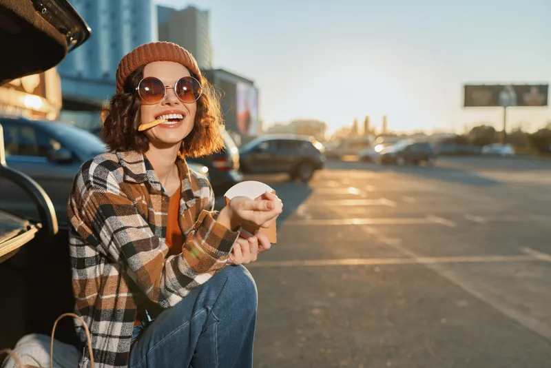 Woman sitting by car in parking lot at sunset, eating a snack with smile and sunglasses, beanie and casual jacket. Authenticity, candid lifestyle and golden hour glow, emotional storytelling.