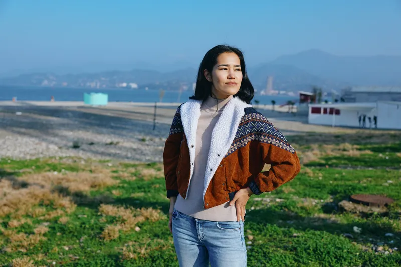 Woman wearing a brown white knit jacket and jeans stands outdoors on a grassy coastal area, confident pose, sunny day, distant mountains, wind swept hair, casual fashion scene