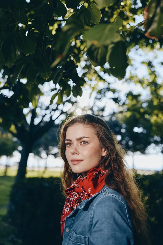 Lifestyle woman in modern boho-western denim street style, wearing red bandana, standing outdoors with soft film color and natural green background in sunny daylight.
