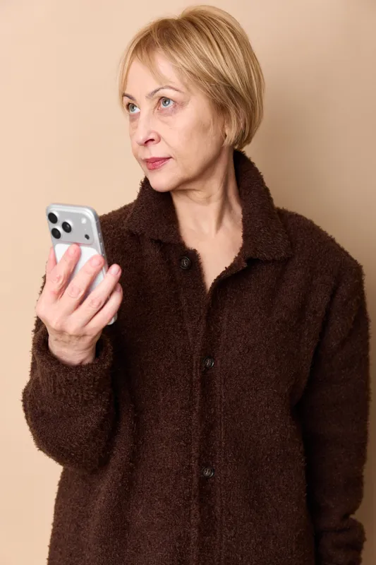 Elderly woman portrait indoors holding smartphone, brown coat, focused gaze, communication and technology, neutral studio background, soft light, gentle