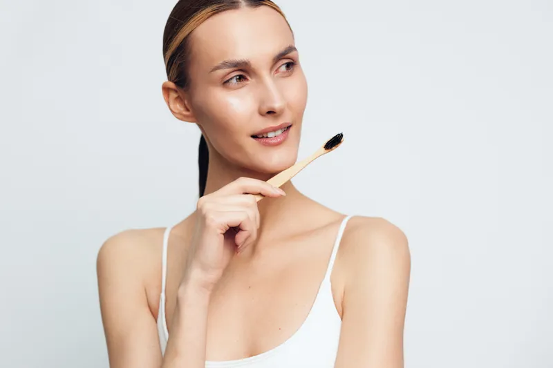 A young woman in a white tank top holds a bamboo toothbrush near her face while looking to the side. The clean background emphasizes dental hygiene and natural beauty care concepts.